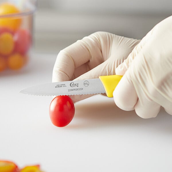 A person in gloves using a Choice serrated paring knife to cut a tomato.