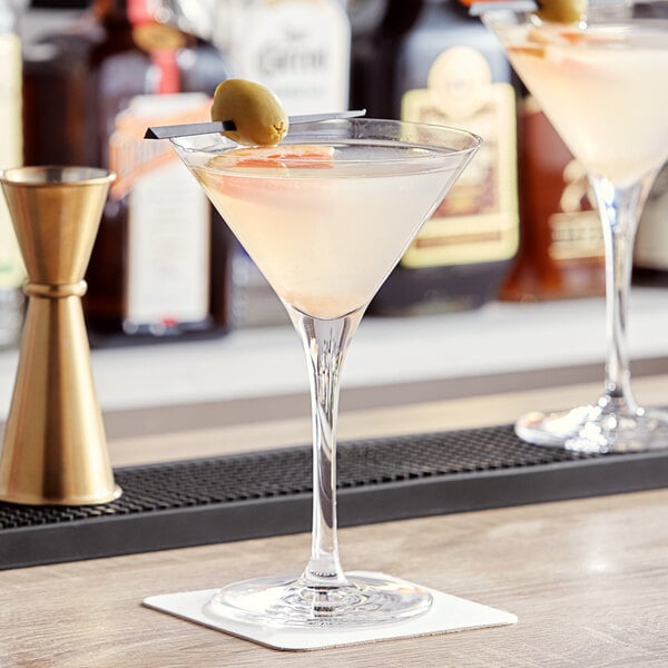 Two Nude martini glasses with green olives on a table.