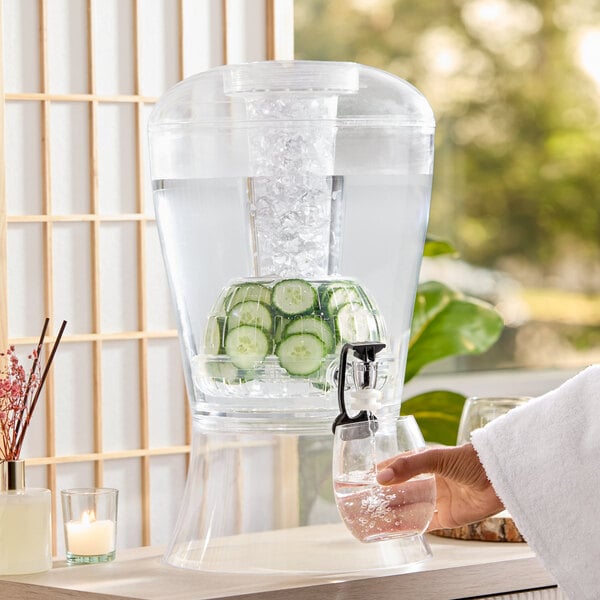 A clear acrylic beverage dispenser with a vertical ice core and fruit infuser, shown filled with water, ice, and cucumber slices.