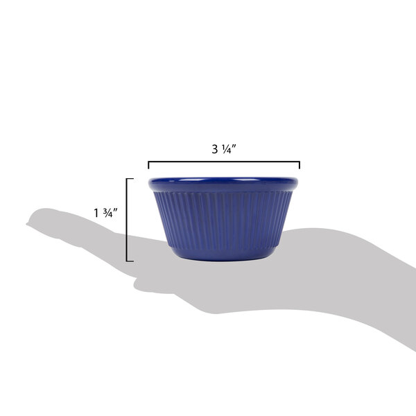 A hand holding a navy blue fluted ramekin with a table in the background.
