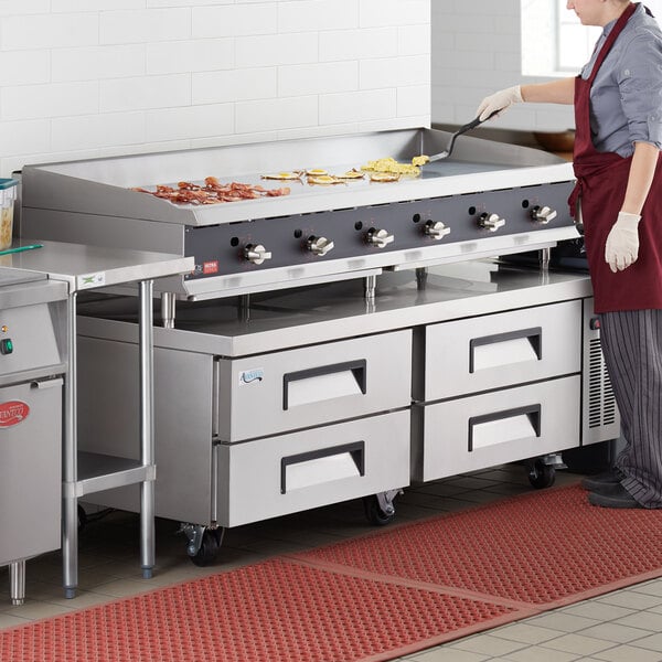 A woman wearing a red apron and gloves cooking on a large stainless steel Cooking Performance Group countertop griddle.