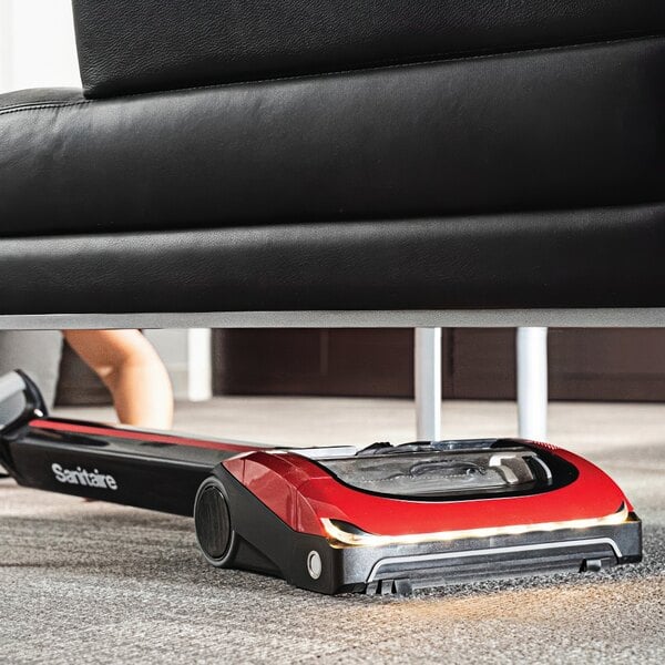 A person using a Sanitaire cordless upright vacuum cleaner to clean under a black leather couch.