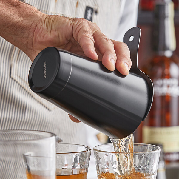 A hand using an Arcoroc Matte Black Stainless Steel Bar Shaker Tin to pour a drink into a glass on a table in a cocktail bar.