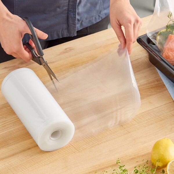 A roll of clear mesh vacuum packaging bags being cut with scissors on a wooden surface.
