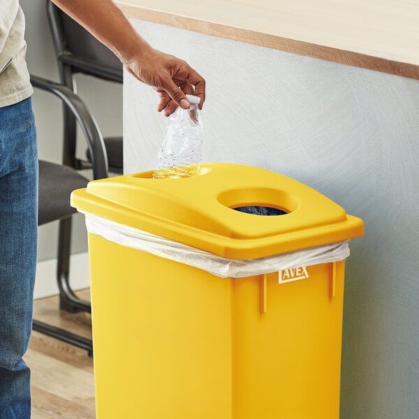 A person putting a plastic bottle into a yellow Lavex Slim Recycling Trash Can with a plastic bag in it.