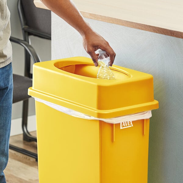 A hand putting a plastic bag into a yellow Lavex slim rectangular trash can.