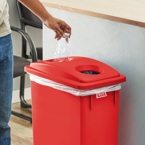 A person putting a plastic bottle into a Lavex red rectangular recycling trash can with a white plastic bag.