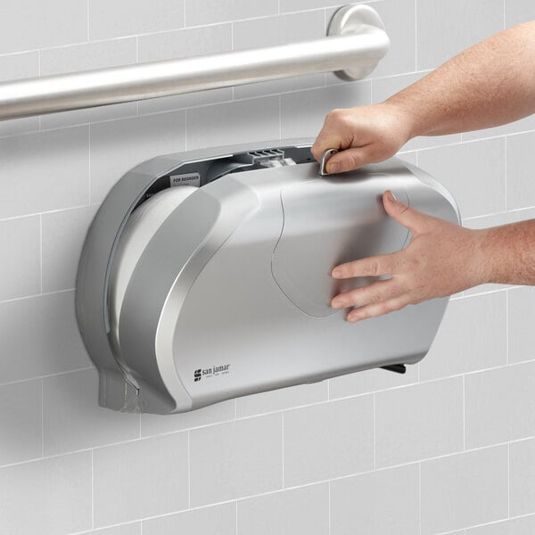 A hand opening a silver San Jamar double roll toilet paper dispenser.