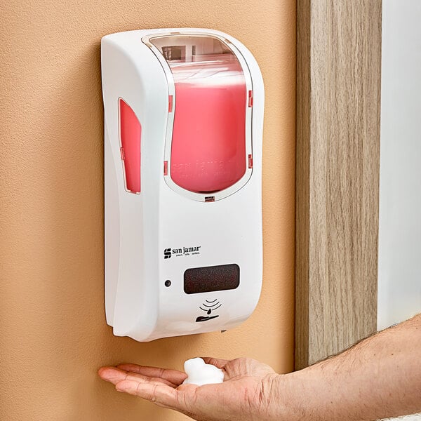 A person using a white San Jamar hybrid automatic foam hand soap and sanitizer dispenser.