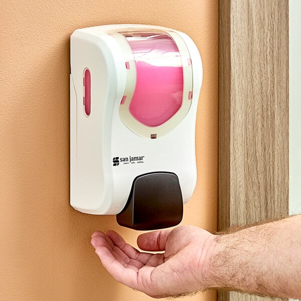 A hand using a white and pink San Jamar manual hand soap, sanitizer, and lotion dispenser.