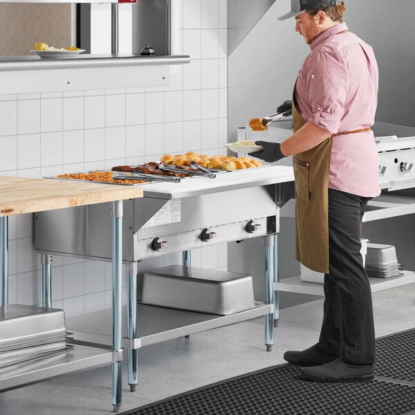 A man in a brown apron using a ServIt liquid propane steam table on a counter in a professional kitchen.