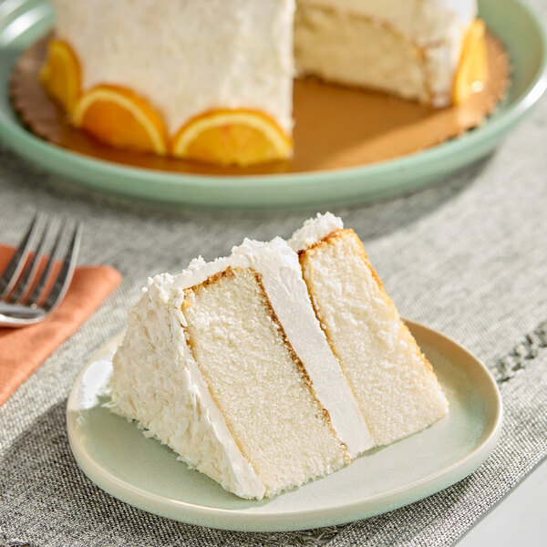 A slice of white layer cake with coconut frosting on a plate, with the rest of the cake in the background.