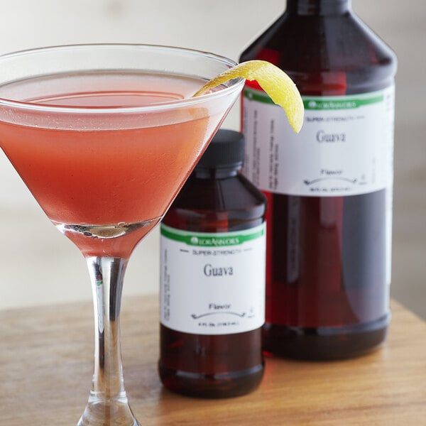 A close-up of a bottle of LorAnn Oils Guava Super Strength Flavor with a glass of pink liquid next to it.