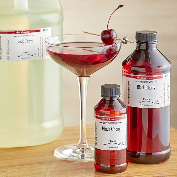 A bottle of LorAnn Oils Black Cherry syrup next to a glass of red liquid with a cherry on top.