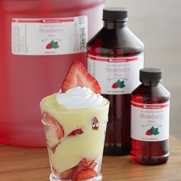 A close up of a glass with a strawberry dessert topped with whipped cream and strawberries with a bottle of LorAnn Oils Strawberry Flavor.