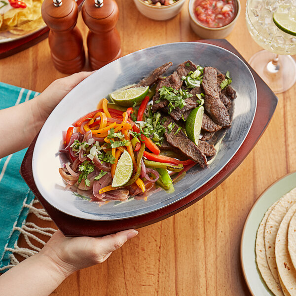 A person holding a Choice oval aluminum sizzler platter with mahogany finish rubberwood underliner full of food.