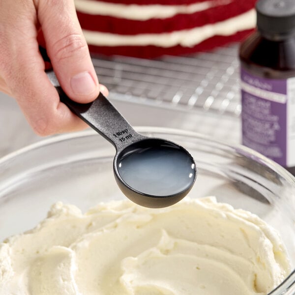 A black measuring spoon filled with liquid being held over a bowl of cream cheese mixture, with a bottle of LorAnn Oils Cream Cheese Bakery Emulsion in the background.