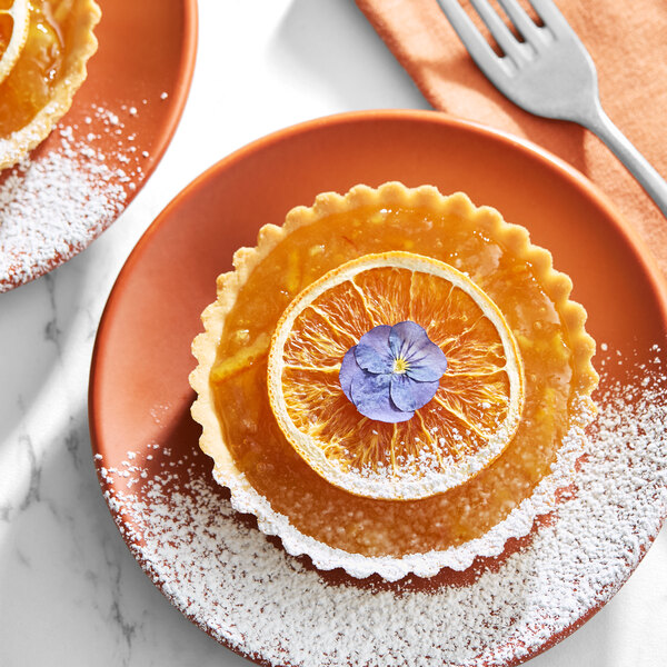 A small tart topped with a slice of orange and a purple edible flower, served on an orange plate with powdered sugar.