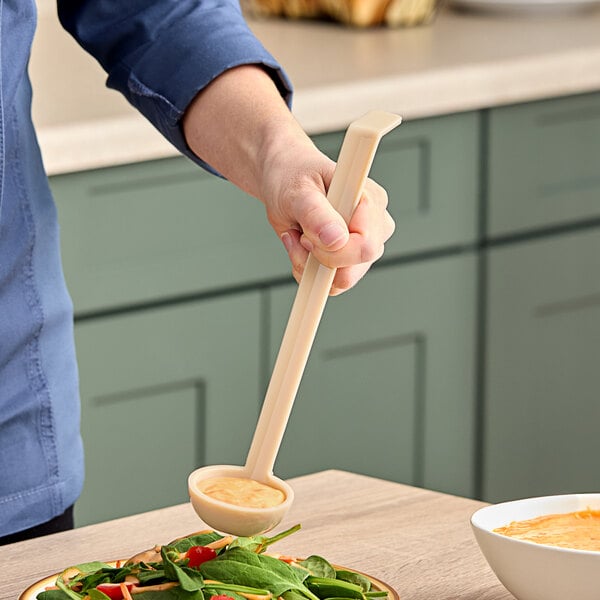 A person using a Cambro beige plastic ladle to scoop sauce from a bowl.