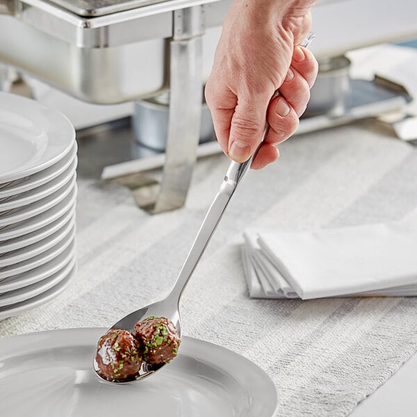 A person holding a Choice stainless steel serving spoon with food on a plate.