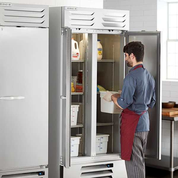 A man in a red apron opening a Beverage-Air reach-in refrigerator with a white container inside.