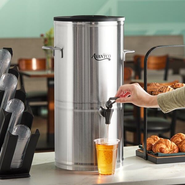 A large stainless steel round iced tea dispenser with a spigot, being used to fill a plastic cup.