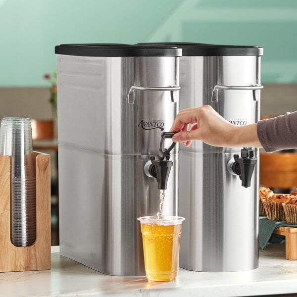A stainless steel 4-gallon slim iced tea dispenser with a stainless steel valve being used to fill a plastic cup.