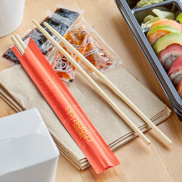 A pair of 9-inch bamboo round chopsticks from Emperor's Select, shown with a red paper sleeve on a stack of napkins.