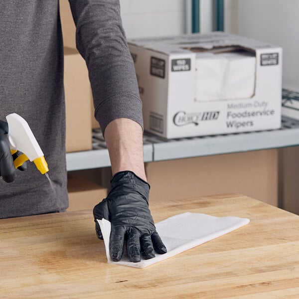 A person wearing a black glove uses a white medium-duty foodservice wiper to clean a wooden surface.