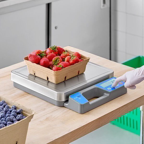 A person weighing a box of strawberries on a grey and blue AvaWeigh digital scale.