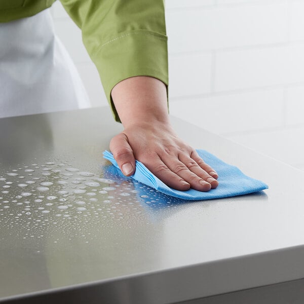 A hand using a Lavex blue general purpose wiper to clean a stainless steel countertop.