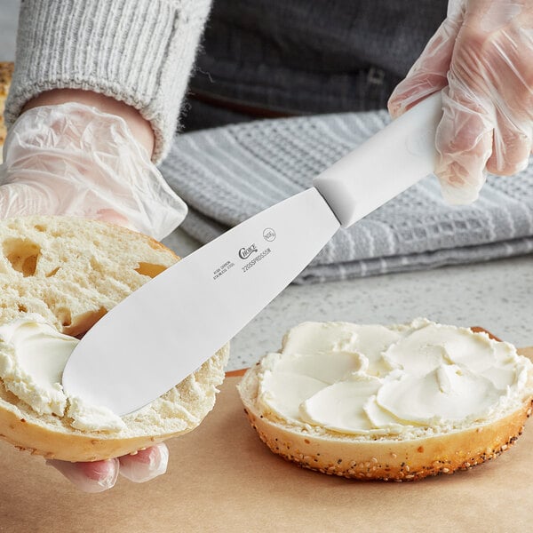 A person using a Choice stainless steel sandwich spreader to spread butter on a bagel.