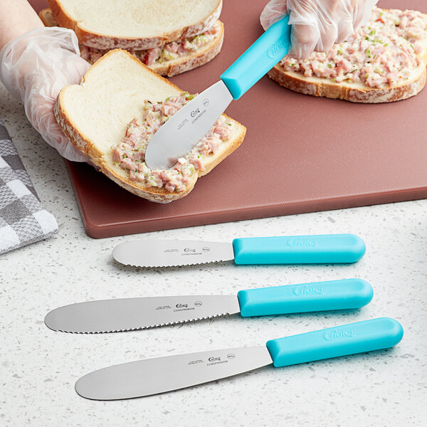 A person using a Choice stainless steel sandwich spreader with a neon blue handle to spread food on a piece of bread.