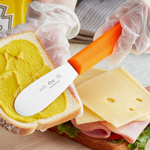 A person using a Choice stainless steel sandwich spreader with a neon orange handle to spread butter on a sandwich.