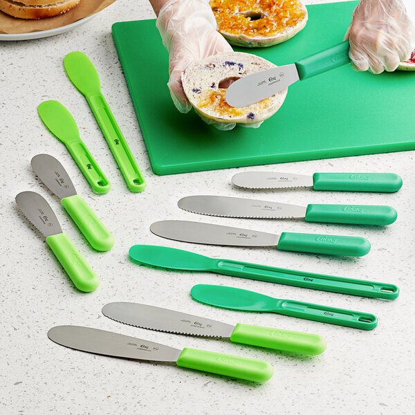 A person using a Choice stainless steel sandwich spreader with a green handle to cut a bagel.