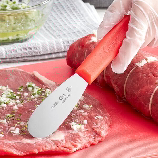 A person using a Choice stainless steel sandwich spreader with a red handle to cut meat.