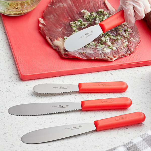 A Choice stainless steel sandwich spreader with a red polypropylene handle being used to spread a food on a cutting board.