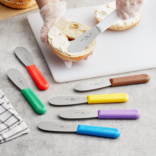 A Choice stainless steel sandwich spreader with a brown handle spreading butter on a bagel.