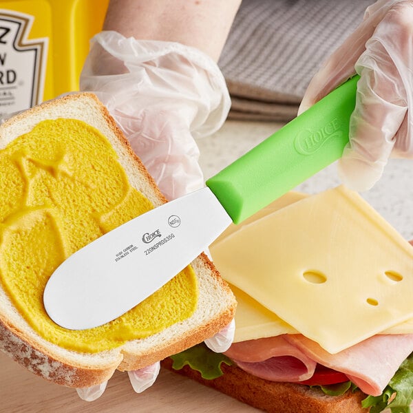 A person using a Choice stainless steel sandwich spreader with a neon green handle to make a sandwich.