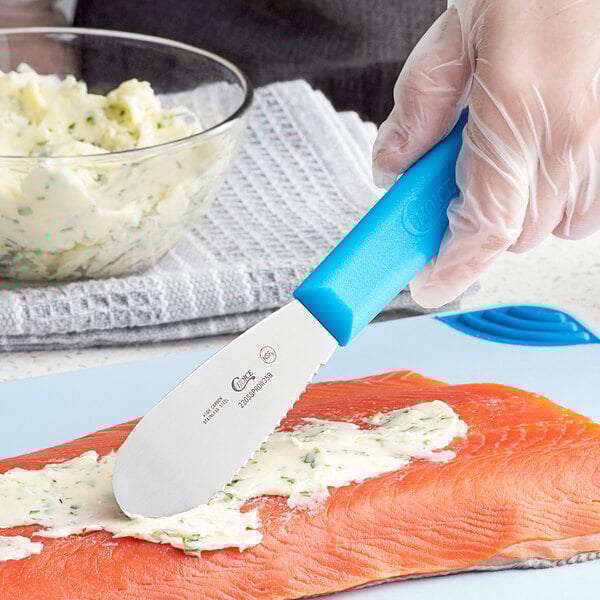 A person's hand with a Choice blue polypropylene handled scalloped sandwich spreader over a piece of food.