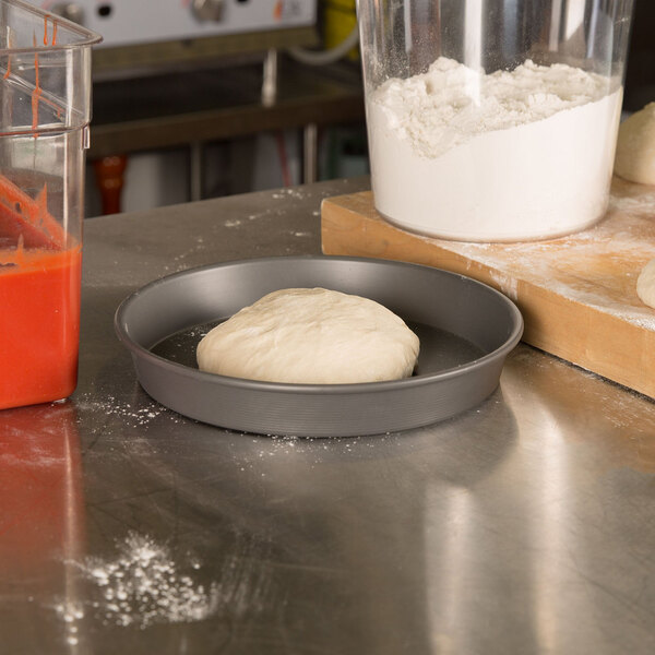 A dough ball in an American Metalcraft Hard Coat Anodized Aluminum Pizza Pan on a counter.