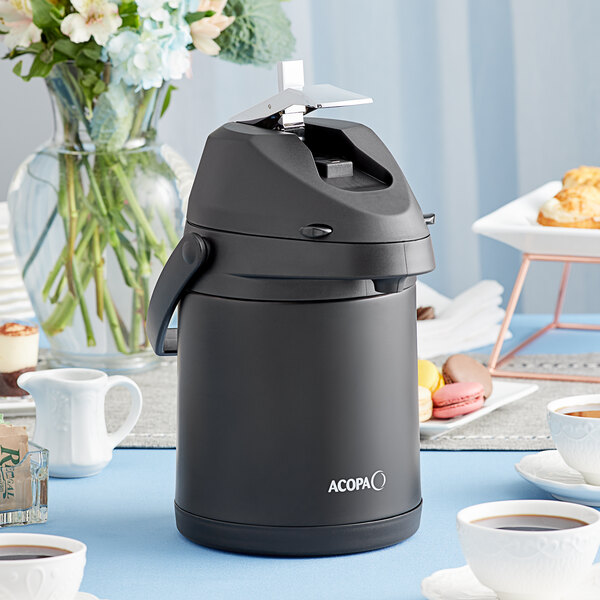 An Acopa matte black stainless steel coffee airpot on a table with a cup of coffee.