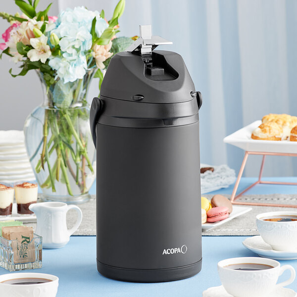 An Acopa matte black stainless steel coffee airpot on a table with a cup of coffee.