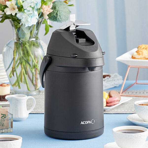 A black Acopa stainless steel coffee airpot with a cup of coffee on a saucer on a table.