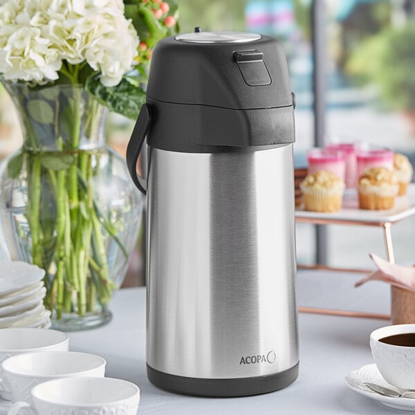 A silver Acopa stainless steel coffee thermos on a table full of coffee cups.