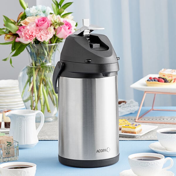 A close-up of a stainless steel Acopa airpot with a metal lever on a table with cups of coffee.