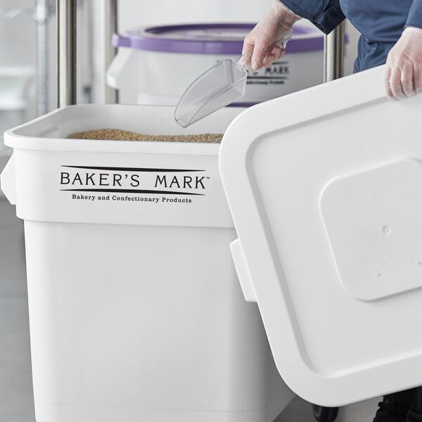 A person using a plastic scoop to fill a Baker's Mark white ingredient storage bin with a lid.