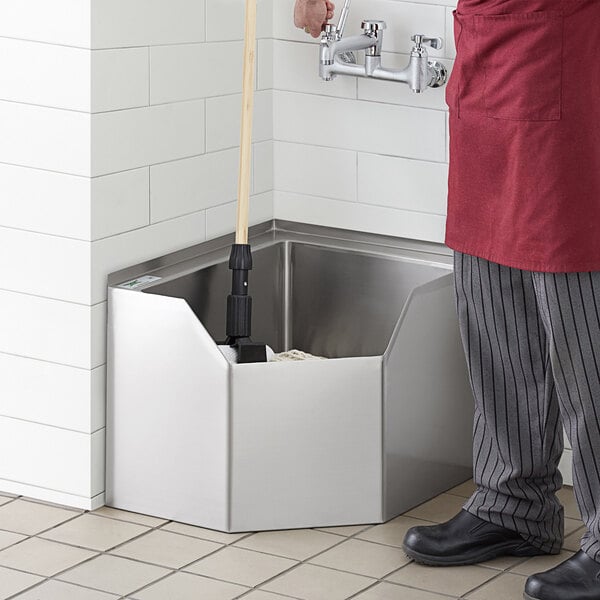 A man in a red apron using a mop to clean a Regency stainless steel corner mop sink.