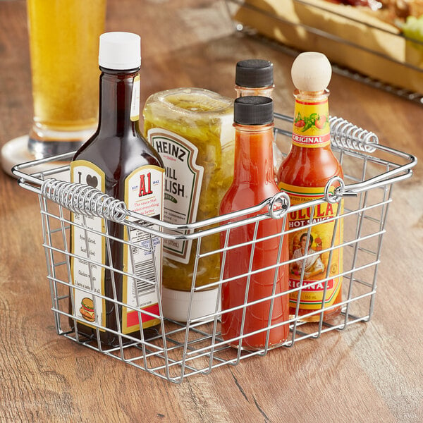 An American Metalcraft chrome market basket filled with bottles of hot sauce on a table.