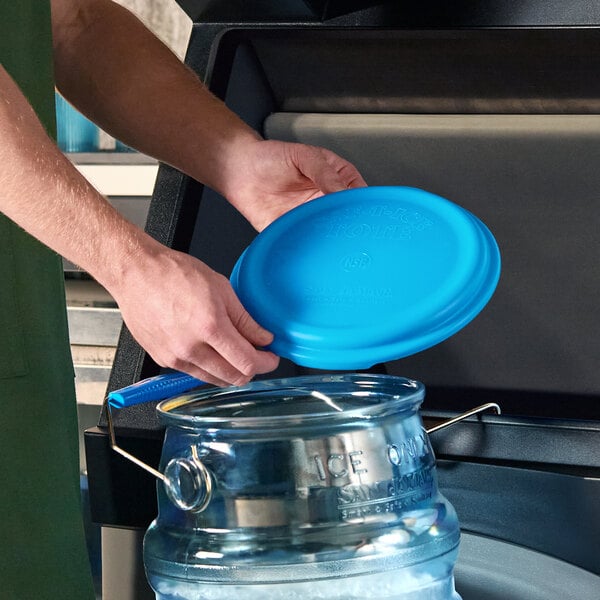 A person is placing a blue lid onto a clear polycarbonate ice tote with metal handles, which is positioned on a hanging bracket.